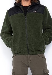 Giacca in pile verde con yoke in nylon nero, chiusura con zip sul davanti, cappuccio, orlo elasticizzato e tasche laterali. Tessuto testurizzato per il calore.