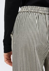 Κοντινό πλάνο ενός ατόμου που φοράει μαύρο και άσπρο παντελόνι με κάθετες ρίγες και ελαστική μέση, καθώς και μαύρη μπλούζα με μακριά μανίκια, με το χέρι χαλαρό στο πλευρό.