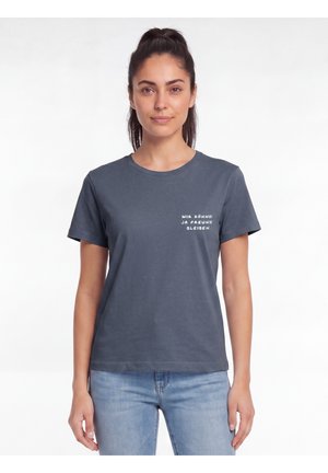 Woman with dark hair tied back wears a gray T-shirt with white German text on chest and light blue jeans, standing against white background.