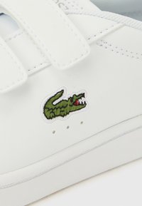 Λευκό δερμάτινο παπούτσι με λουράκια με κλείσιμο hook-and-loop και πράσινο λογότυπο κροκοδείλου. Διαθέτει ραμμένες διακοσμήσεις και μικρές οπές στη σόλα.