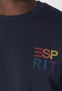 Námořnicky modrá mikina s žebrovaným výstřihem; obsahuje vícebarevný vyšitý text "ESP РIT" v červené, oranžové, žluté, zelené, modré a fialové barvě.