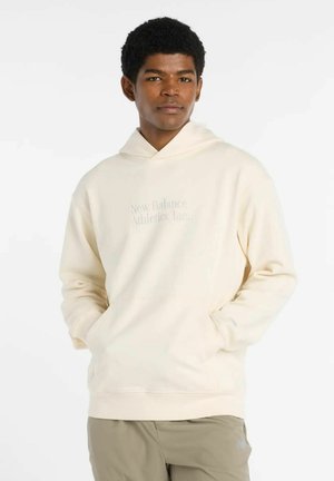 Young man wearing a cream New Balance hoodie with hands in pockets and beige pants, standing against a plain white background.