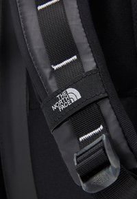 Czarny pasek od plecaka z logo The North Face, wykonany z teksturowanej tkaniny, z białymi paskami i plastikową klamrą do regulacji.
