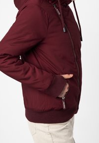 Burgundy zip-jacka med huva, ribbade ärmslut och nederkant. Har två sidofickor, slät yta och dragsko för justerbar passform.
