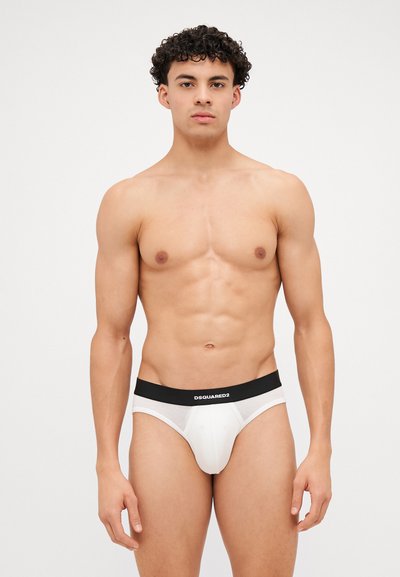 Slips pour hommes blancs avec une ceinture noire arborant le logo "DSQUARED2". Le tissu semble lisse et extensible, avec une conception ajustée.