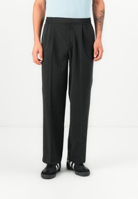 Pantalones gris oscuro con un ajuste relajado, cinturilla elástica y una textura sutil. Combinados con zapatillas negras que tienen rayas blancas.