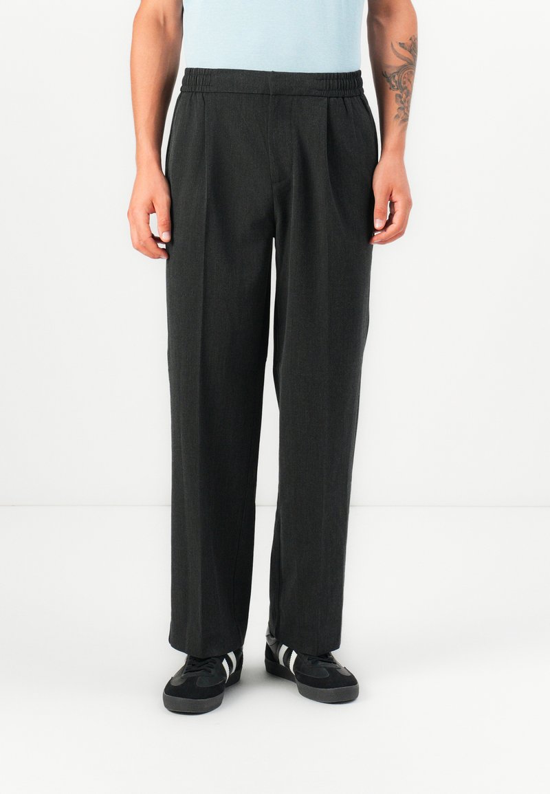 Pantalones gris oscuro con un ajuste relajado, cinturilla elástica y una textura sutil. Combinados con zapatillas negras que tienen rayas blancas.