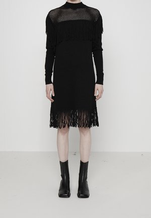 Schwarzes Strickkleid mit einem Mesh-Oberteil und Fransenverzierungen an den Schultern und dem Saum, kombiniert mit knöchelhohen schwarzen Stiefeln.