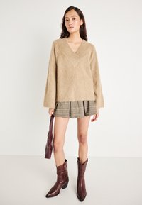 Mulher vestindo um suéter beige de V com textura felpuda, saia mini plissada xadrez, botas de cowboy borgonha, segurando uma bolsa borgonha combinando ao seu lado.