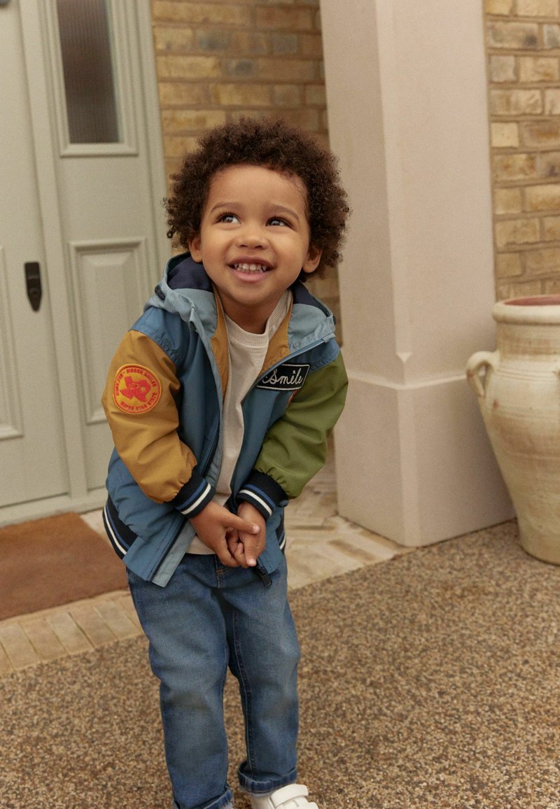 Veste colorée avec des panneaux bleus, verts et bruns, ornée d'un patch souriant. Associée à une chemise blanche et un jean bleu, se tenant sur un sol texturé.