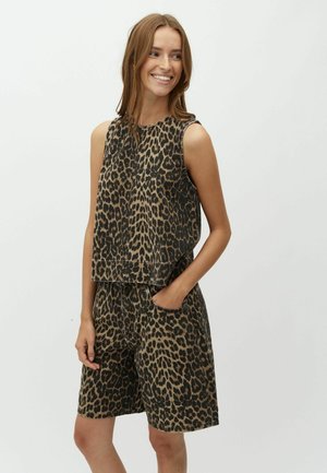 Uśmiechnięta kobieta, ubrana w bezrękawnik z nadrukiem w lamparta oraz pasujące szorty, stoi z jedną ręką w kieszeni na jednolitym tle.