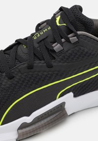 Scarpa sportiva nera con tomaia in rete, accenti giallo-verdi a contrasto, lacci grigi e un dettaglio laterale testurizzato visibile.