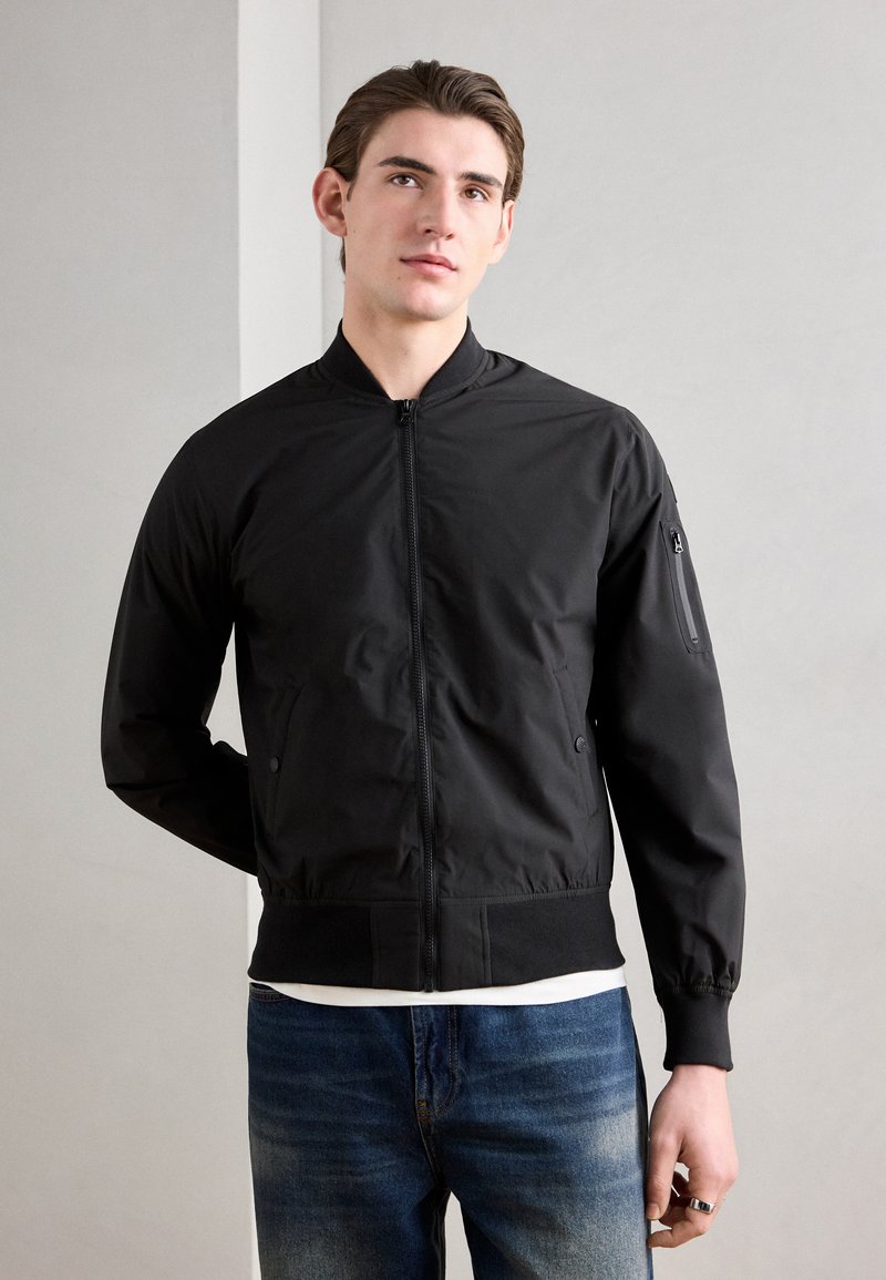 Schwarze Bomberjacke mit Reißverschluss auf der Vorderseite, geripptem Saum und Bündchen; verfügt über Seitentaschen und eine Reißverschlusstasche am Ärmel. Getragen mit blauen Jeans.