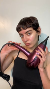 Suszarka do włosów w intensywnym bordowym odcieniu z czarną dyszą, wyposażona w teksturowane uchwyty, przycisk zasilania oraz długi przymocowany kabel.