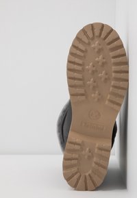 Semelle d'une botte beige avec des crampons profonds comportant des motifs en croix et le logo Timberland, placée droite contre une surface blanche.