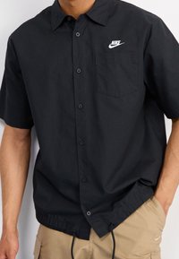 Černá košile s krátkým rukávem na knoflíky, vyrobená z lehkého materiálu, se malým bílým logem Nike na levé náprsní kapse a nastavitelným šňůrkovým lemem.