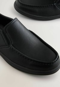 Black slip-on shoes made of textured leather. Rounded toe with stitched detailing and rubber sole for grip. Streamlined, minimal design.