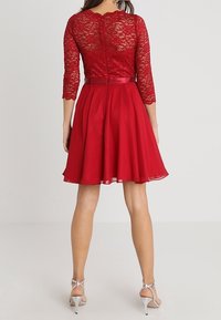 Robe rouge avec des manches en dentelle trois-quarts, corsage ajusté et jupe évasée. Ceinture en satin et fermeture éclair au dos. Texture lisse.