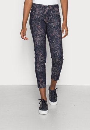 Femme portant un pantalon ajusté, longueur cheville, de couleur sombre avec un motif floral subtil, et des baskets noires à lacets avec des semelles blanches.