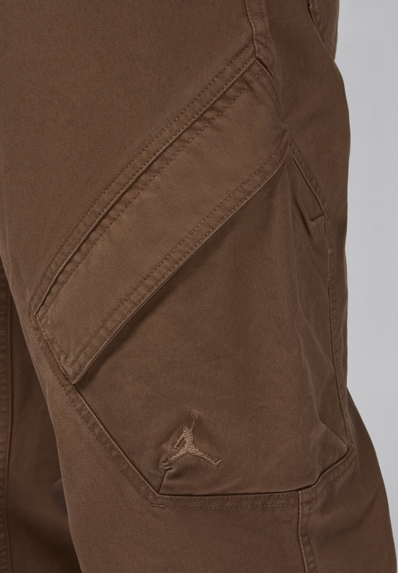 Pantaloni cargo marroni realizzati in tessuto resistente, con tasca a patch e logo Jumpman ricamato, caratterizzati da una texture liscia e un design sartoriale.