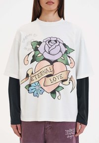 Witte oversized t-shirt met een grafisch ontwerp waarop een paarse roos, een hart en de tekst "ETERNAL LOVE" staan, gecombineerd met een zwarte longsleeve eronder.