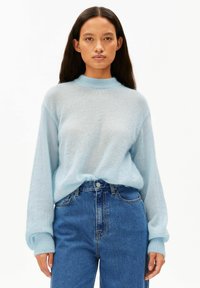 Hellblauer, halbtransparenter Strickpullover mit geripptem Ausschnitt und langen Ärmeln. Getragen zu hoch taillierten blauen Jeans.