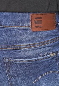 Granatowe dżinsy z ciemnego denimu z brązową skórzaną metką z napisem "G-STAR RAW." Wyraźne przeszycia oraz zaokrąglone tylne kieszenie podkreślają design.