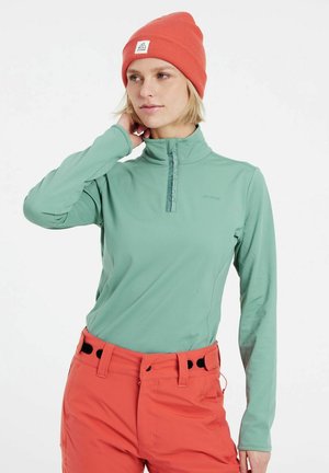 Lichtgroene shirt met lange mouwen, hoge kraag en een gedeeltelijke rits, gecombineerd met feloranje broek met zwarte accenten en zijzakken.