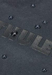 Zwarte textuurstof met waterdruppels en een reliëf "THULE" logo, met een duurzame en waterafstotende ontwerp.