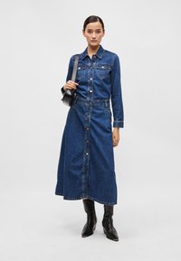 Mørkeblå denimkjole med lange ærmer, knappelukking foran og to brystlommer. Brukt med svarte ankelstøvler.