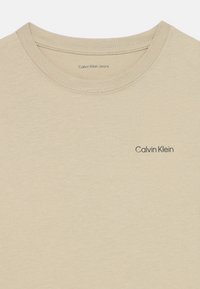 Béžové bavlnené tričko s okrúhlym výstrihom a krátkymi rukávmi. Na prednej strane sa nachádza malé čierne logo "Calvin Klein Jeans". Hladká textúra.