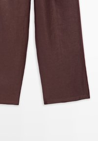 Pantalon brun droit avec coutures visibles aux ourlets sur un fond clair uni.