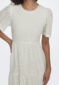 Valkoinen eyelet-mekko, jossa on pyöreä pääntie, lyhyet puff-hihat, chevron-kuvio ja kerroksellinen hame. Sisältää herkkiä leikkauksia hengittävyyden parantamiseksi.