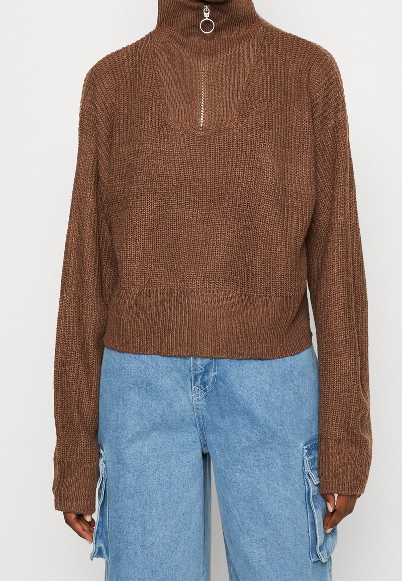 Pull en tricot marron à col roulé avec fermeture éclair argentée associé à un pantalon cargo en denim bleu clair.