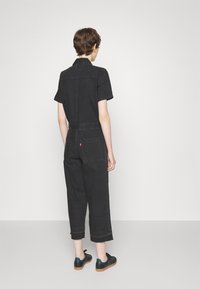 Svart denimoverall med korta ärmar, avskurna ben och bakfickor. Har en figursydd midja och kontrasterande sömmar. Bärs med svarta sneakers.