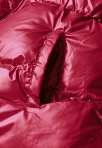 Rød dynejakke i close-up, med skinnende, quiltet stof, synlig lynlås og teksturale folder. Inkluderer en åben lomme detalje.