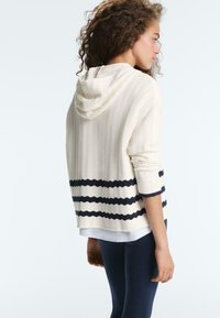Femme aux cheveux bouclés portant un sweat à capuche crème texturé avec des rayures noires sur les manches et l'ourlet, pantalon bleu marine, vue de profil sur fond blanc.