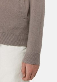 Ljusbrun stickad tröja med en jämn yta, prydd med ribbade muddarna och en ficka på sidan, visad mot en vit bakgrund.