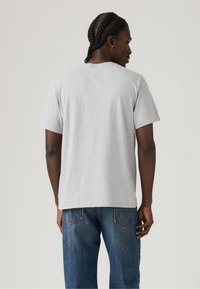 Man met gevlochten haar die een eenvoudig lichtgrijze T-shirt met korte mouwen en blauwe jeans draagt, van achteren gefotografeerd tegen een neutrale achtergrond.