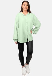 Camicia button-up di un verde chiaro, con texture e maniche lunghe, abbinata a leggings neri lucidi e sneakers bianche. Design di un outfit casual.