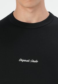 Black short-sleeve T-shirt with a ribbed neckline and white embroidered "Originals Studio" logo on the chest. Cotton material, smooth texture.