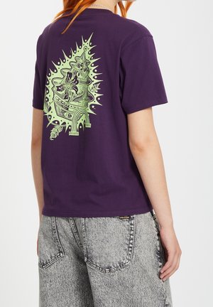 T-shirt violet arborant un motif graphique vert complexe au dos, représentant un mélange de fleurs et de formes abstraites. Manches courtes.
