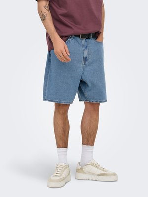 Hombre con pantalones cortos de mezclilla azul holgados, calcetines blancos, zapatillas beige y una camiseta granate con brazos tatuados sobre un fondo liso.