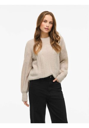 Pull en tricot beige clair avec un motif texturé, doté d'un col rond et de manches tombantes. Associé à un jean noir taille haute.
