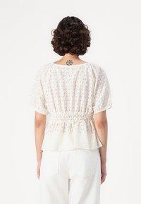 Blouse blanche texturée avec des manches courtes et bouffantes et une taille froncée. Motif ajouré sur l'ensemble, créant une apparence légère et aérée.