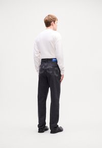 Ung mand vender væk iført en hvid langærmet skjorte, sorte jeans med "HUGO" label på bagsiden af taljen, og sorte sko på hvid baggrund.
