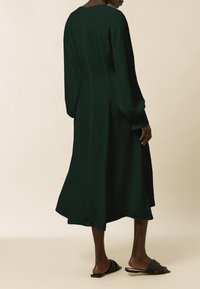 Vestido de manga larga, color verde oscuro, con un dobladillo fluido, textura suave y escote redondo; combinado con sandalias negras de tipo sin hebillas.