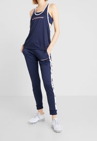 Canotta sportiva blu navy con rifiniture bianche e logo, abbinata a pantaloni jogger blu navy con strisce laterali bianche e logo, completata da sneaker bianche.