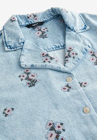 Cămașă din denim bleu deschis cu broderie florală, având guler, închidere cu nasturi și o textură moale. Accentele includ detalii în roz și verde.