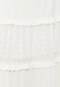 Tissu blanc avec deux coutures horizontales froncées et une section centrale en dentelle florale transparente ornée de petites fleurs brodées.